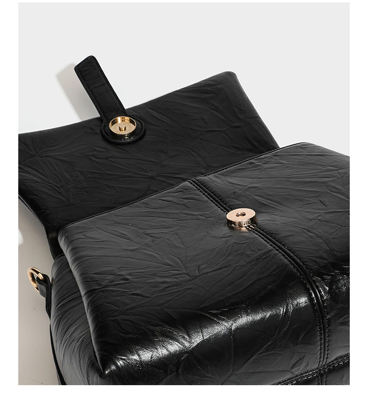 Structured black satchel bag with glossy crinkled leather-look finish, gold-tone emblem flap closure, slim top handle, and