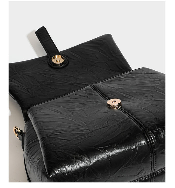 Structured black satchel bag with glossy crinkled leather-look finish, gold-tone emblem flap closure, slim top handle, and